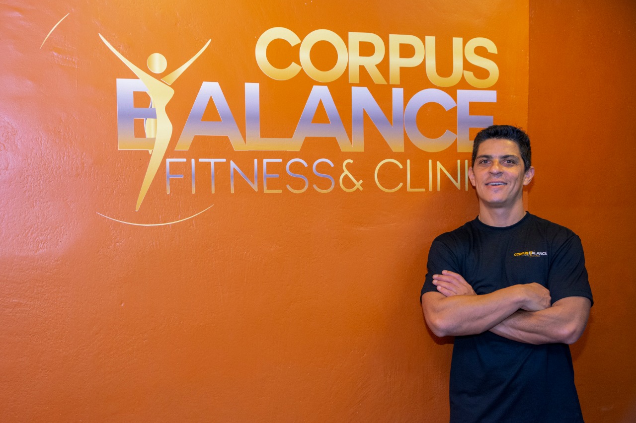 Dalton Capita de braços cruzados ao lado do logotipo da Corpus Balance Fitness & Clinic na parede laranja